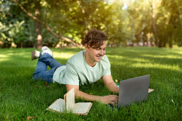 Cheerful student guy doing homework with laptop and book, resting in university campus outdoors, lying on lawn