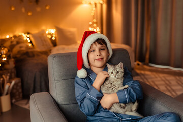 Obraz premium a 10-year-old boy in blue pajamas and a red Christmas cap is holding a small British kitten in his hands. decorated room with magical christmas lights