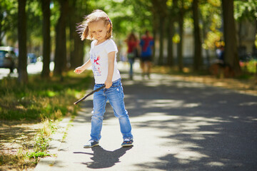 Preschooler girl playing with a stick on a sunny street. Outdoor summer activities for kids. Active games for children
