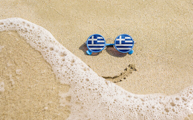 Fototapeta na wymiar Sunglasses with Greek flags on a sandy beach. Nearby is a sea lightning and a painted smile. The concept of a successful vacation in the resorts of Greece.