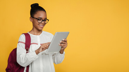 Cheerful smart teen afro american girl pupil in glasses with backpack studying with digital tablet,...