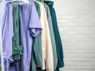 Lilac and green t-shirt, sweatpants, sneakers, sweatshirt. Women's, youth clothes of different colors hang on a hanger. White brick wall.