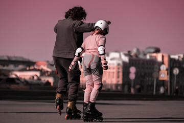 A male coach teaches a child to roller-skate in the city. A girl in a helmet and fall protection is...