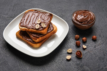 Plate of bread with chocolate paste and hazelnuts on black background