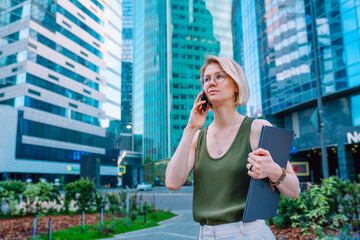 Young blonde businesswoman with laptop making call by phone outdoors against skyscrapers. Business lady in glasses speaking on smartphone while walking through financial district. Selective focus