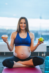 Healthy and junk food. Athletic smiling pregnant woman in sport top sit on purple yoga mat in gym and make choice between donut and green apple. Healthy lifestyle and healthcare concept. 