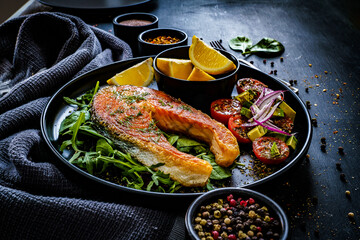 Fried salmon steak with potatoes and fresh vegetable salad served on black plate on wooden table
