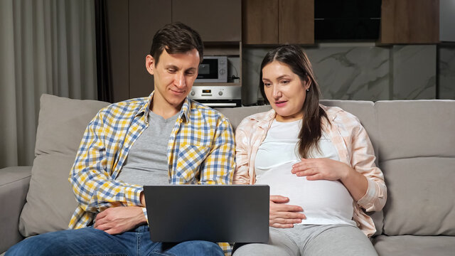 Brunet husband and pregnant wife smile doing online shopping for future baby via laptop choosing stroller and crib sitting on sofa in living room.