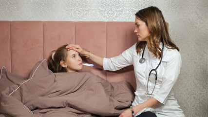 Fototapeta premium Lady doctor in white robe measures temperature putting thermometer into mouth of sick teen girl lying on bed covered with blanket in bedroom at home.