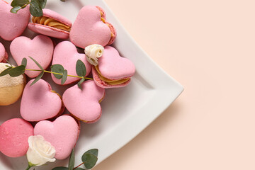 Plate with tasty heart-shaped macaroons and flowers on beige background