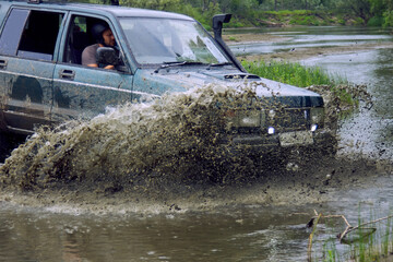 An SUV or off-road vehicle crosses a river with splashes of dirty water. A helmeted driver crosses...