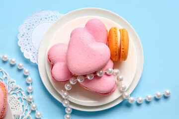 Plate with tasty heart-shaped macaroons and beads on blue background