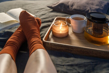 Soft photo of woman`s legs in the woolen socks on the bed with book and cup of tea and candle on...