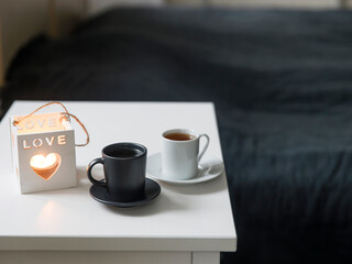 Fototapeta na wymiar A two cups of coffee: white and black on a tray and a candlestick with the inscription love is on the bedside table