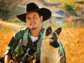 Front view photo of a cowboy sitting on a yellow field with two dogs. Amazing rural image of a real farmer wearing a classic cowboy black hat with his dogs in the countryside of Brazil 
