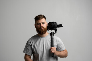 Fototapeta na wymiar Portraite of bearded cinematographer in a grey t-shitr with a camera and 3-axes gimbal on a white background. Videography, Filmmaking, hobby and creativity concept.