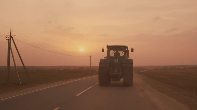 Farm tractor with large wheels driving on country road at sunset after sowing or harvesting in field.