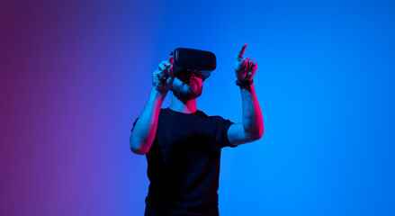 Young man uses virtual reality headset. Cheerful bearded man in t-shirt moving hands and using VR headset in neaon light background. Technology concept.