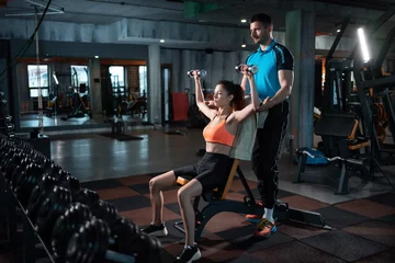Tuinposter young female exercising in gym with dumbbell with personal athletic trainer © goami