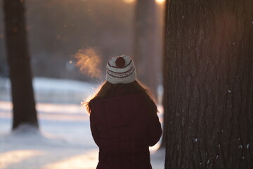 woman in a warm hat and jacket during winter looking out to the sunset at a wooded park while breathing in the cold air