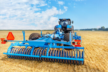 Fototapeta premium A powerful universal blue harrow on an agricultural tractor against the background of a harvested yellow wheat field.