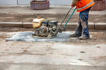 A worker with a petrol vibrating plate patches a pothole on an old road.