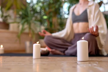 Foto op Plexiglas Cropped young healthy woman is practicing yoga at home sitting in lotus pose on yoga mat meditating relaxed alone, keeping calm. Mindfulness meditation concept.focus on candles, side view © Roman