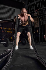 Fototapeta na wymiar Sport. Battle ropes session. Attractive young fit caucasian sportsman working out in functional training gym doing exercise with battle ropes. In sports club. Portrait, single shirtless male