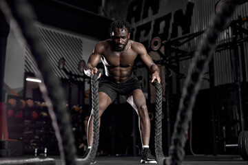 afro american fit sportsman doing battle ropes exercise at crossfit gym. African Man wear black shorts training with rope. sport motivation, cross fit, fitness concept. cardio training