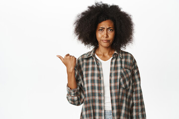 Skeptical african american female model, woman 30 years old frowning disapproval, pointing left, complaining, dislike smth, white background