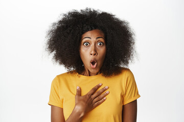 Fototapeta na wymiar Enthusiastic Black woman looking surprised, staring impressed and fascinated at camera, standing in yellow t-shirt over white background