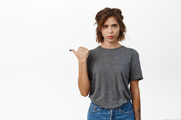 Sad and disappointed young woman pointing finger left, looking upset and uneasy, regret, showing...