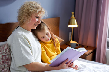 nice grandmother reads a book to little daughter lying on bed in cozy children's room, domestic atmosphere. friendly aged woman enjoys spending time with grandchildren, relaxing together