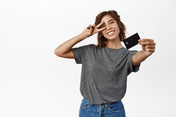 Fototapeta premium Portrait of beautiful young woman showing credit card, recommending bank, contactless shopping or discounts in store, time to shop, standing over white background