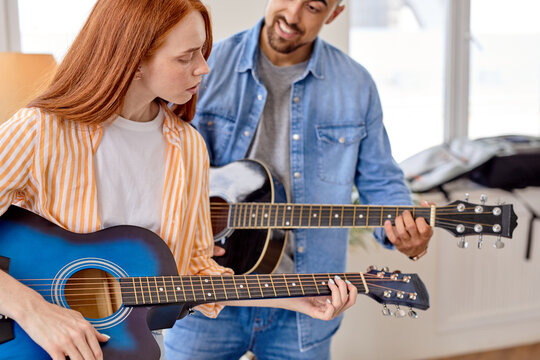 Smiling man in casual wear teaching confident girlfriend to play the guitar. Guy shows woman how to play guitar. Couple in casual wear using musical instrument
