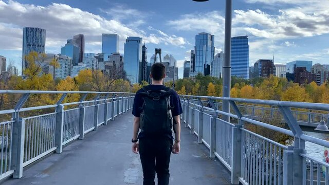 Young Man walking across Bow river bridge towards city center. Man walks towards skyscrapers in Calgary city. Canada