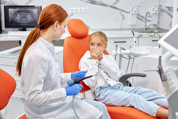 Pretty caucasian child girl sit on couch looking frightened by dental drill in hands of professional female dentist, in modern bright clinic. beautiful little child is closing mouth by fear, scared