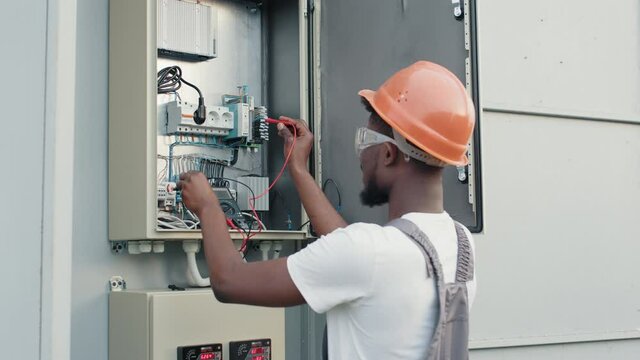 African american electrician in uniform and helmet standing near box with cables and doing service work on solar station. Maintenance, people and green energy concept. Technician at solar farm.