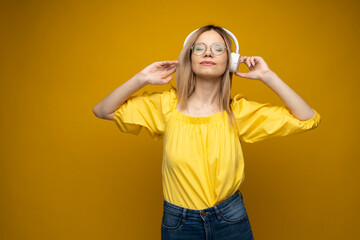 Beautiful attractive young blond woman wearing yellow t-shirt and glasses in white headphones listening music, dancing and laughing on blue background in studio. Relaxing and enjoying. Lifestyle.