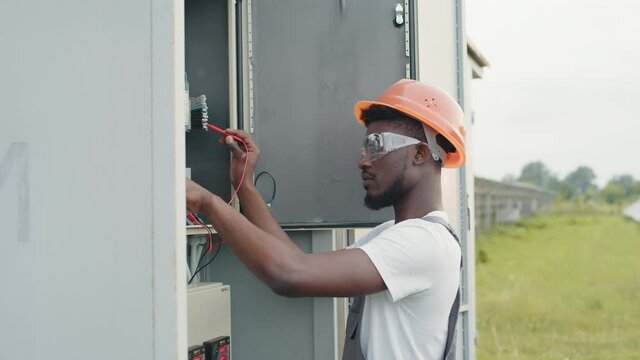 Competent technician in orange helmet and grey overalls standing near switchgear and connection solar station to common network. Production of alternative energy and saving of environment