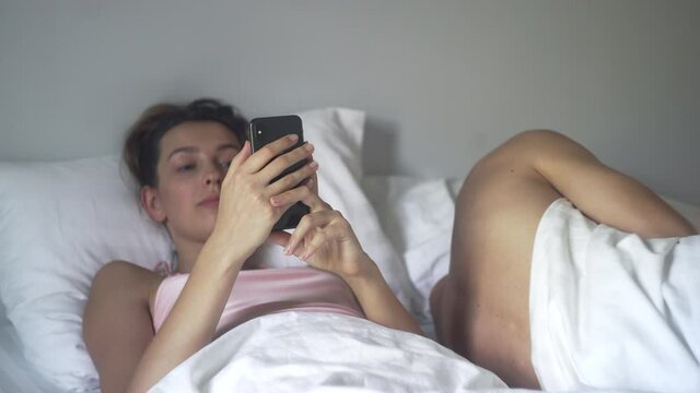 Young Caucasian woman scrolling her phone in bed in the morning. Night or morning using phone in bed concept. Phone addict problem.