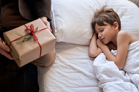 Christmas morning concept. man in caot put present on bed next to lying kid in white bed.happy moments in winter holidays. cropped father celebratin sleepy son, at home. happy new year