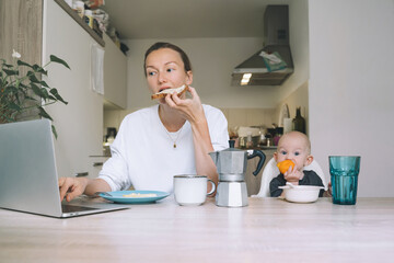 Young woman studying or working online at home while having breakfast with her baby on kitchen....