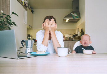 Young mother with coffee fighting tiredness while breakfast with baby in kitchen. Freelancer mom and child after sleepless night. Woman studying or working online at home on maternity leave