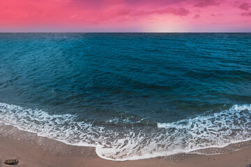 Vibrant saturated seascape with white waves rolling in on the beach with a rock, turquoise-colored seawater, and pink foggy clouds at sundown under the horizon.