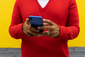 Adult male holding a blue cell phone with both hands. He is wearing a red sweater and white flannel. Yellow and gray wall background. Technology concept.