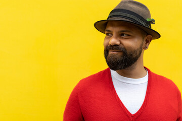 Adult Latino male (39) with beard, looks to his right side with an optimistic gesture. He is wearing a hat, red sweater and white flannel. Yellow wall background. Copy space. Body language concept