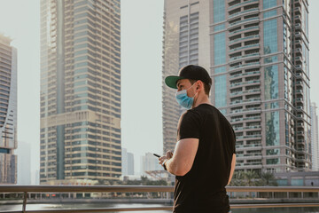 Person tourist with medical mask holding and using  phone by typing messages while standing outdoor on the street with urban background and skyscrapers at sunset time