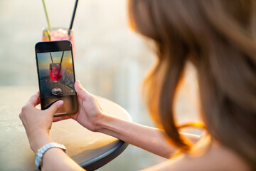 Confidence Asian woman using mobile phone photographing tasty cocktail drink on the table while waiting for meeting with friends at skyscraper outdoor rooftop restaurant in the city  at summer sunset
