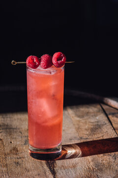 pink cocktail in tall Collins glass with ice and three raspberries on cocktail pick
